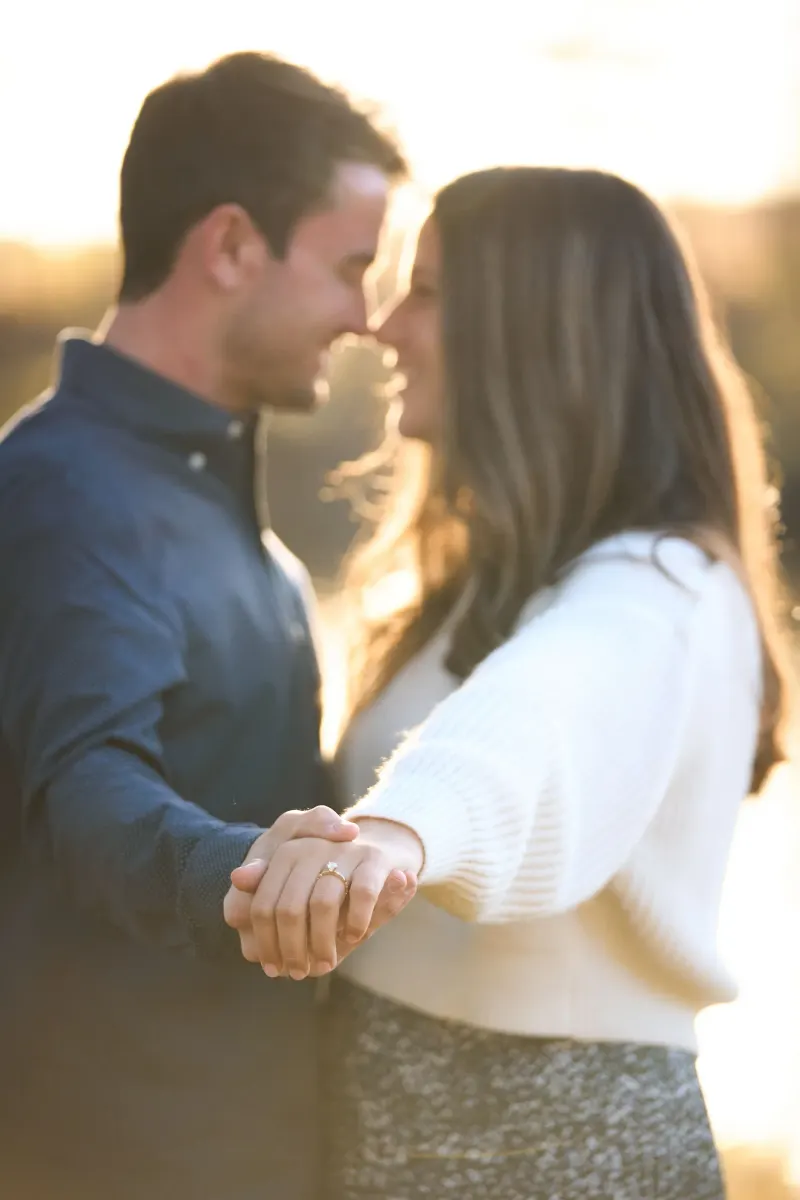 A couple holding hands and about to kiss during golden hour outdoors, with the man wearing a blue shirt and the woman in a white top displaying a ring on her hand.