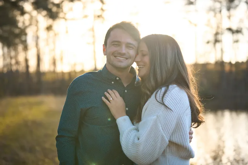 A smiling couple embraces outdoors during golden hour near a calm pond, with pine trees lining the background and warm sunlight filtering through the forest.