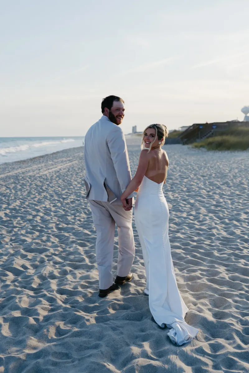 A bride in a white wedding dress and groom in a light linen suit hold hands on a sandy beach at golden hour, with ocean waves and coastal cliffs visible in the background.