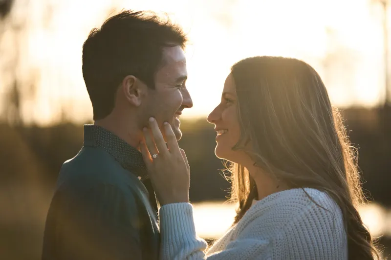 A man and woman face each other closely during golden hour, with the man touching his face while the woman smiles at him beside a body of water.